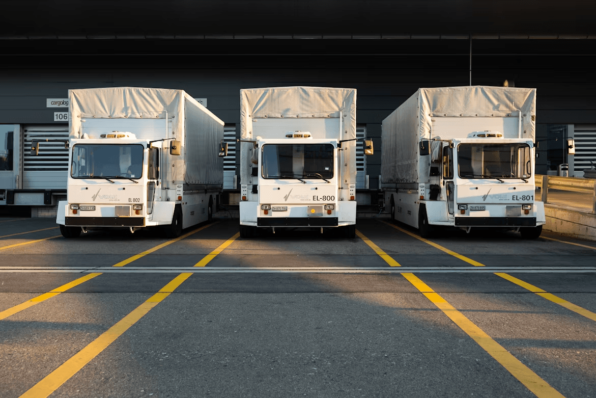Three white delivery trucks with canvas-covered cargo boxes, labeled EL-800, EL-801, and EL-802, parked side by side at loading docks, photographed head-on in warm late-day light with yellow lane markings on the pavement.