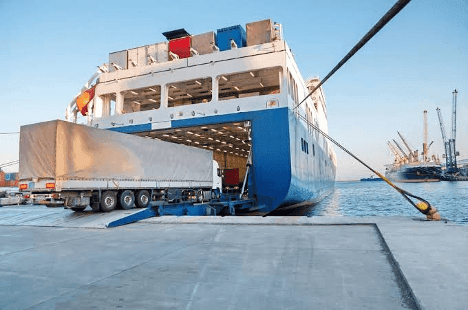 A roll-on/roll-off (RoRo) cargo ship docked at port with its bow ramp open, connected to a truck trailer for loading/unloading