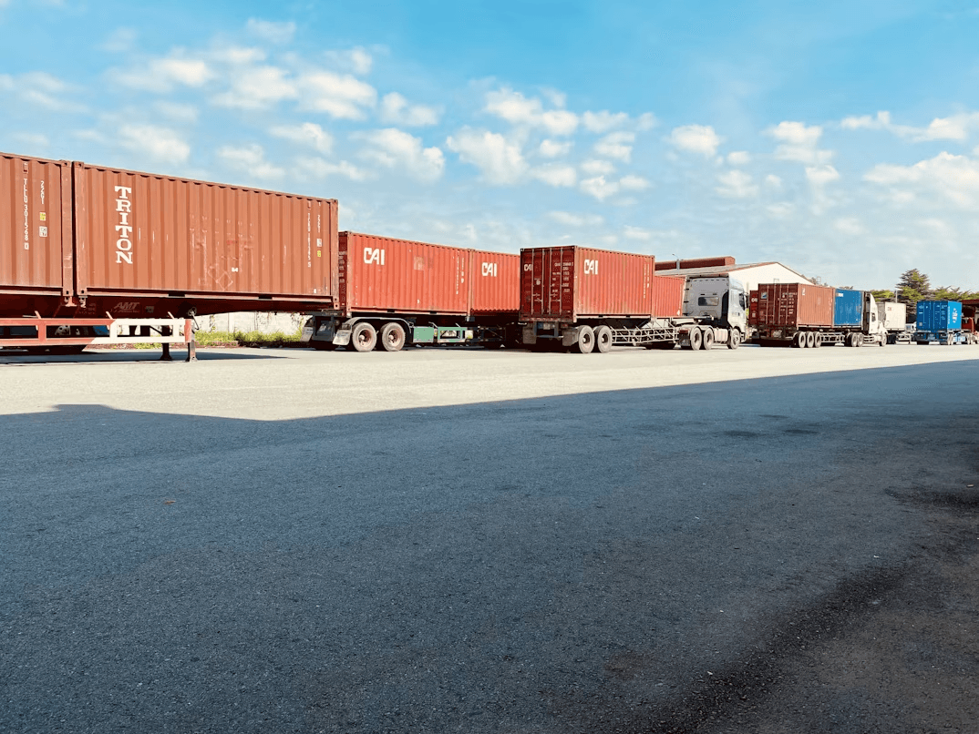Row of flatbed trucks carrying shipping containers labeled TRITON and CAI, parked in a line at a shipping yard under a partly cloudy blue sky.
