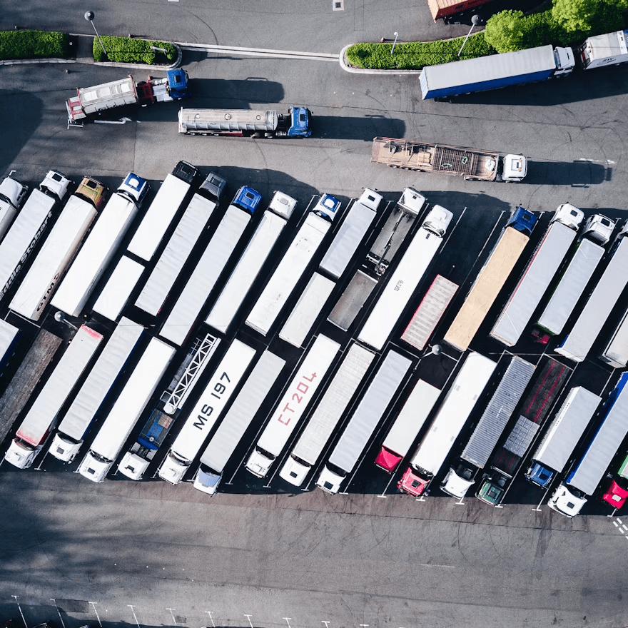 Aerial view of a truck parking lot with rows of semi-trailers parked diagonally in marked bays, their white and colored roofs creating a striped pattern; a few trucks maneuver in the open lane at the top near a grassy roundabout.